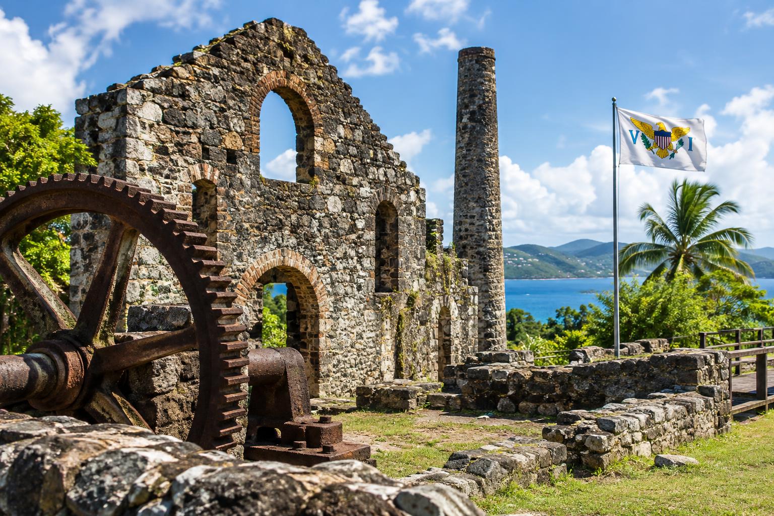 Weathered stone ruins of a historic Caribbean sugar mill under bright daylight, with the US Virgin Islands flag visible in the background, emphasizing the educational and historical significance of the site.
